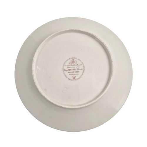 fig 3. A circular white porcelain plate featuring a gilded rim, a red and pink floral border, and a central armorial crest of a bird beneath a coronet with a banner reading STRIKE.