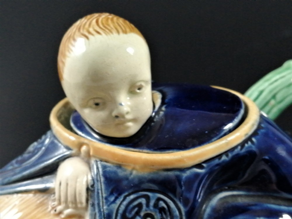 A Victorian earthenware majolica teapot shaped as a melon with a figure of a boy in a blue jacket seated on top forming the lid. View X