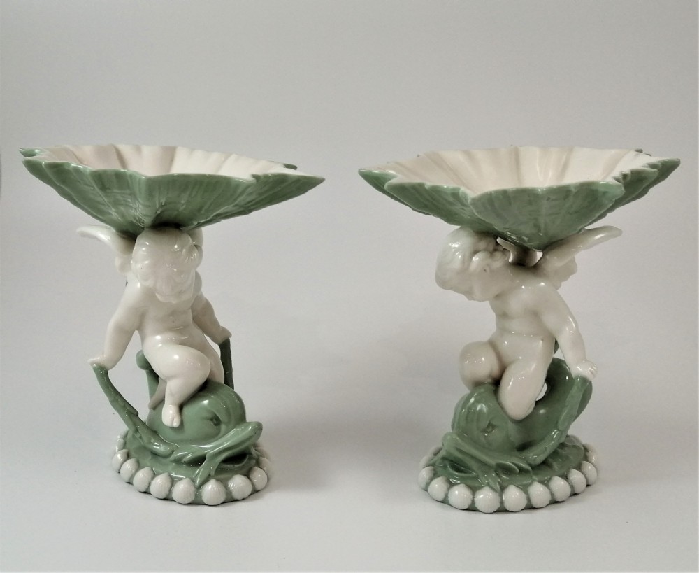 A pair of Victorian ceramic compotes featuring unglazed white cherub figures holding green leaf-shaped bowls aloft while seated on green glazed dolphin bases.