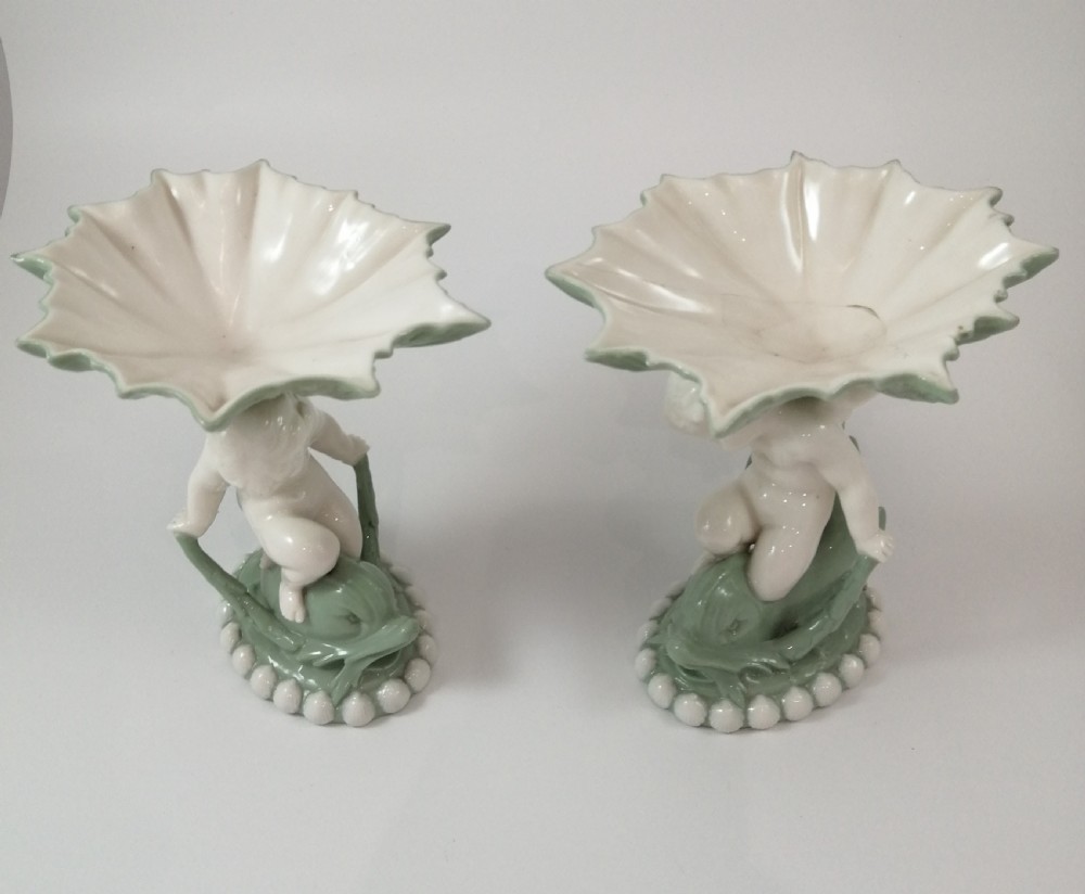 A pair of Victorian ceramic compotes featuring unglazed white cherub figures holding green leaf-shaped bowls aloft while seated on green glazed dolphin bases. View II