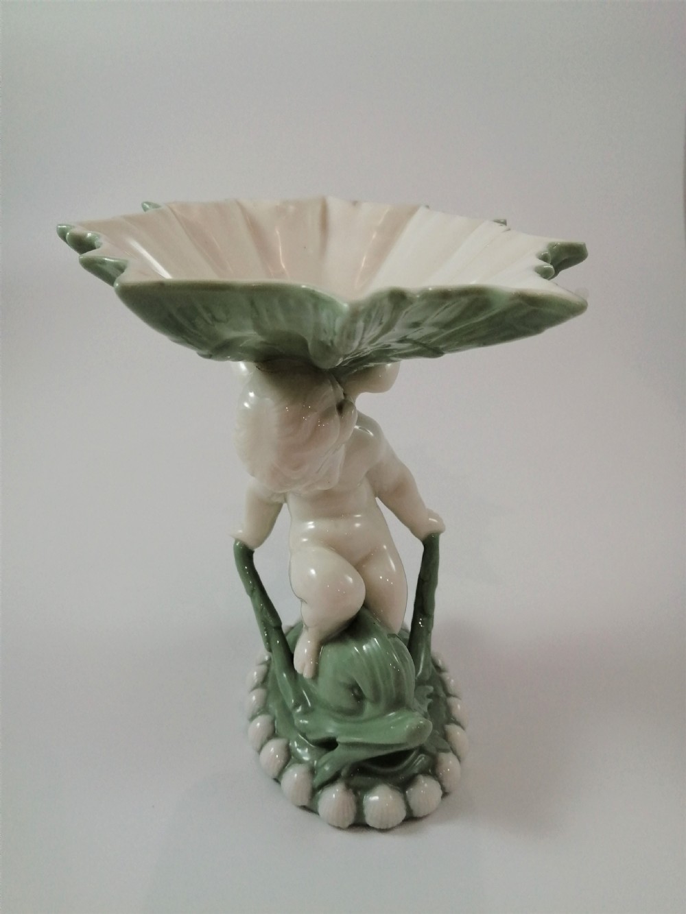 A pair of Victorian ceramic compotes featuring unglazed white cherub figures holding green leaf-shaped bowls aloft while seated on green glazed dolphin bases. View XII