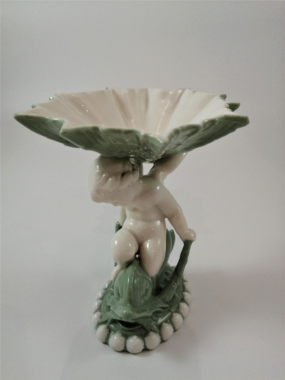 A pair of Victorian ceramic compotes featuring unglazed white cherub figures holding green leaf-shaped bowls aloft while seated on green glazed dolphin bases. View XVI