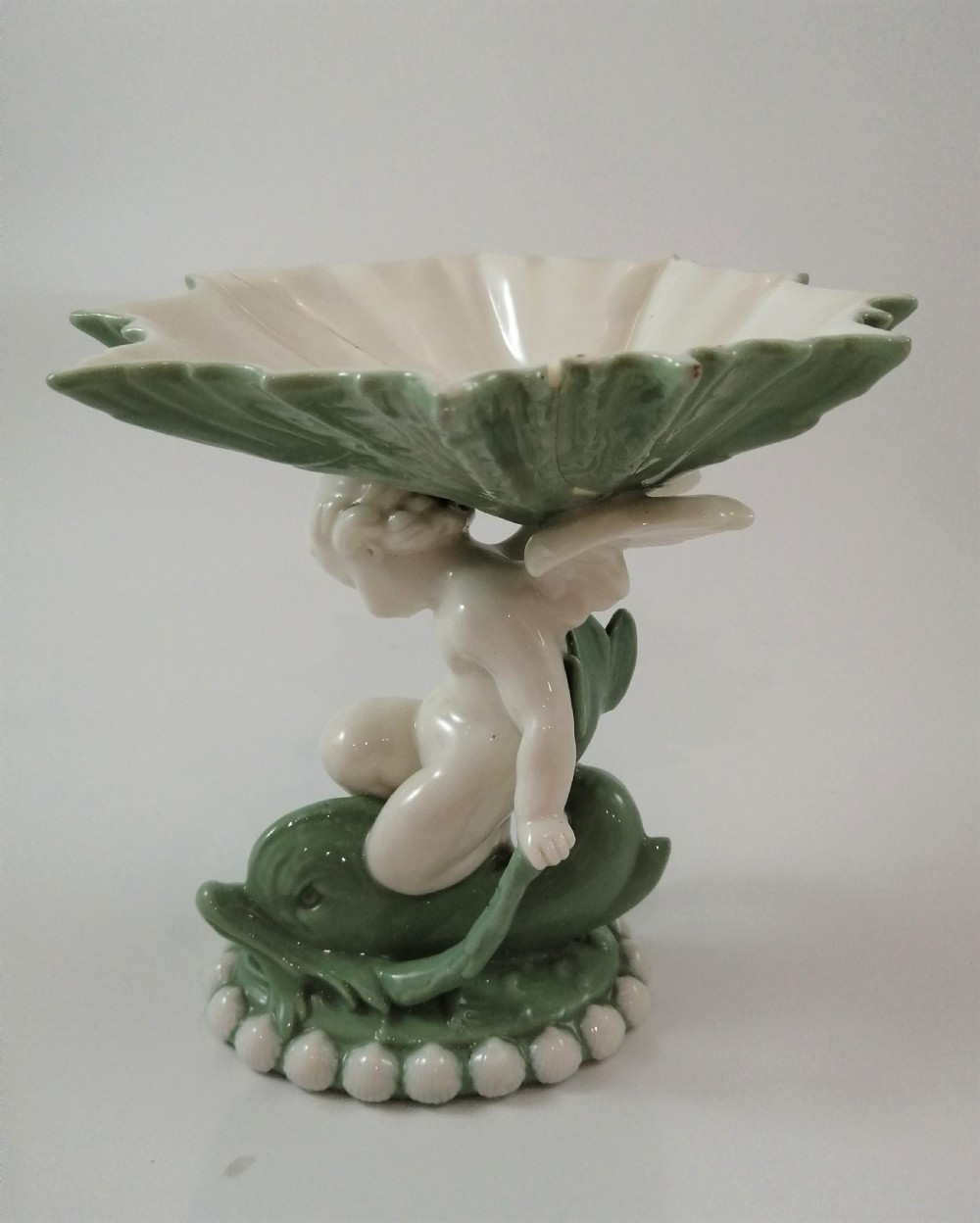 A pair of Victorian ceramic compotes featuring unglazed white cherub figures holding green leaf-shaped bowls aloft while seated on green glazed dolphin bases. View XVII