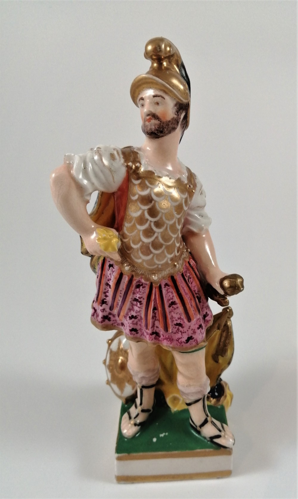 An antique hand-painted porcelain figure depicting the Roman god Mars in gilded armour, a pink tunic, and a golden helmet, standing on a green base.