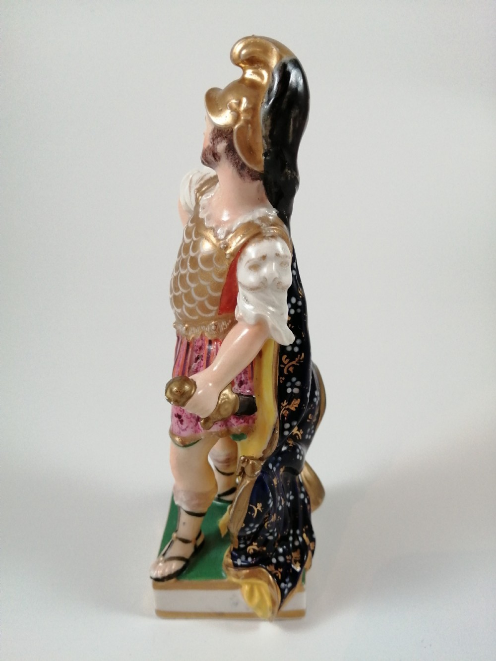 An antique hand-painted porcelain figure depicting the Roman god Mars in gilded armour, a pink tunic, and a golden helmet, standing on a green base. View III
