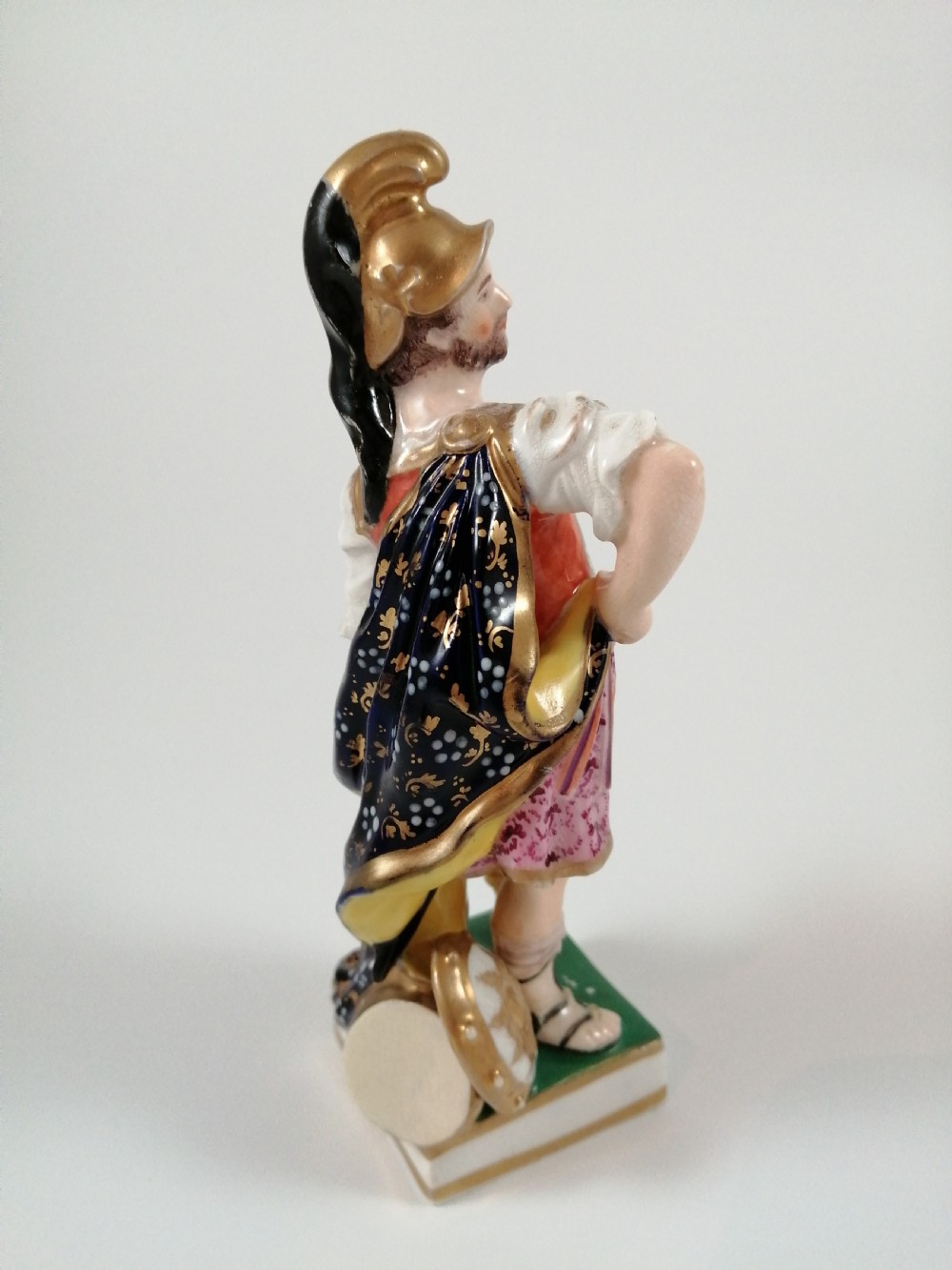 An antique hand-painted porcelain figure depicting the Roman god Mars in gilded armour, a pink tunic, and a golden helmet, standing on a green base. View VI