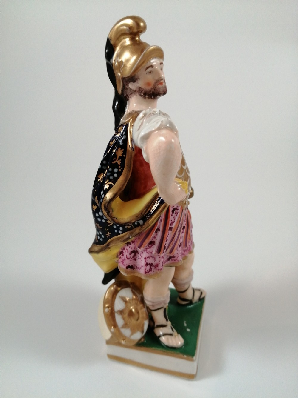 An antique hand-painted porcelain figure depicting the Roman god Mars in gilded armour, a pink tunic, and a golden helmet, standing on a green base. View VII