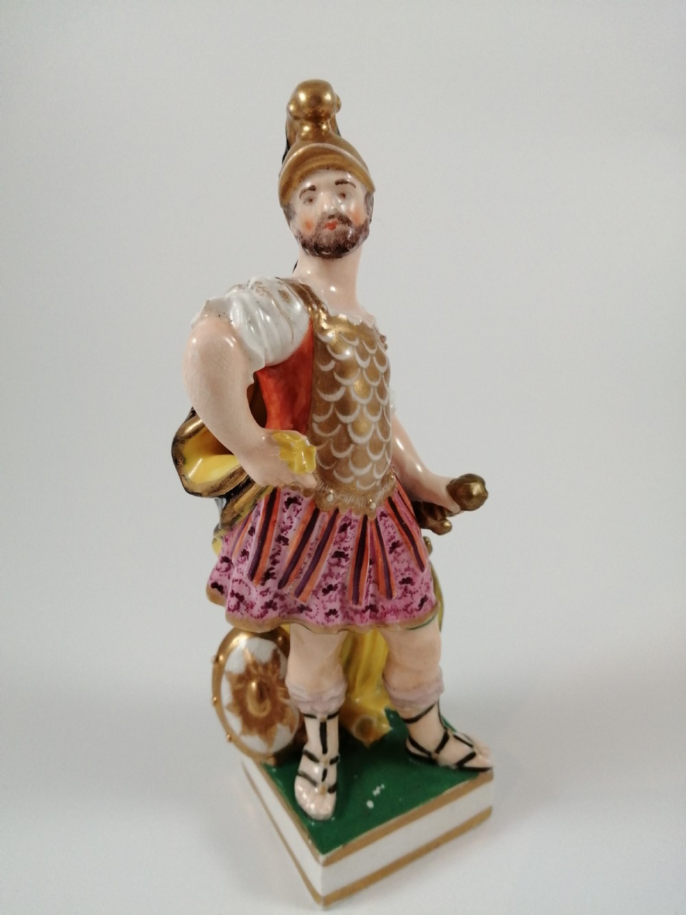 An antique hand-painted porcelain figure depicting the Roman god Mars in gilded armour, a pink tunic, and a golden helmet, standing on a green base. View VIII