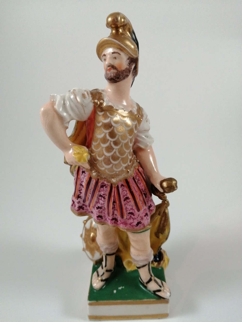 An antique hand-painted porcelain figure depicting the Roman god Mars in gilded armour, a pink tunic, and a golden helmet, standing on a green base. View IX