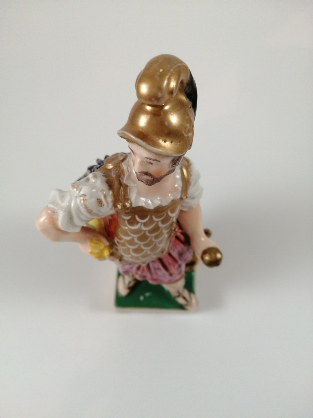 An antique hand-painted porcelain figure depicting the Roman god Mars in gilded armour, a pink tunic, and a golden helmet, standing on a green base. View X