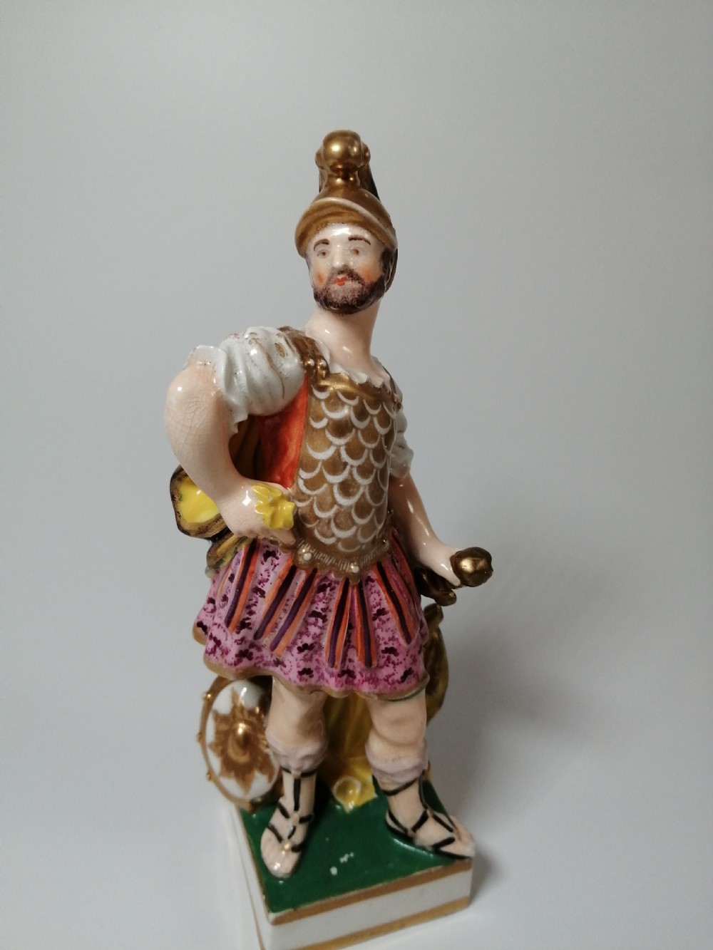 An antique hand-painted porcelain figure depicting the Roman god Mars in gilded armour, a pink tunic, and a golden helmet, standing on a green base. View XI