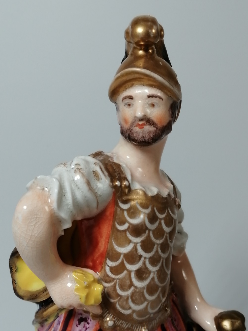 An antique hand-painted porcelain figure depicting the Roman god Mars in gilded armour, a pink tunic, and a golden helmet, standing on a green base. View XII