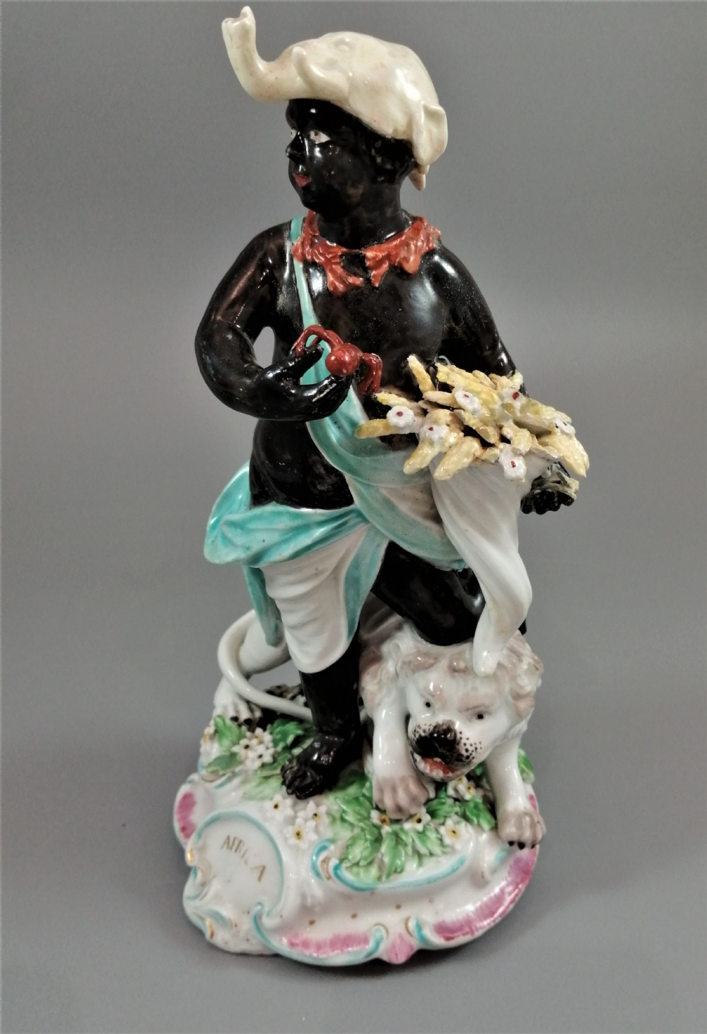 An 18th-century Derby soft-paste porcelain figurine depicting an allegorical figure of Africa wearing an elephant scalp headdress beside a crouching lion on a rococo scroll base.