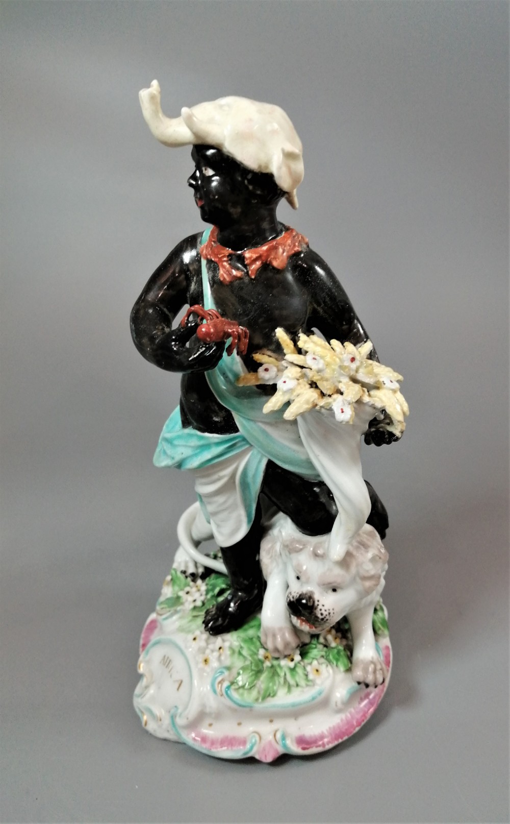 An 18th-century Derby soft-paste porcelain figurine depicting an allegorical figure of Africa wearing an elephant scalp headdress beside a crouching lion on a rococo scroll base. View III