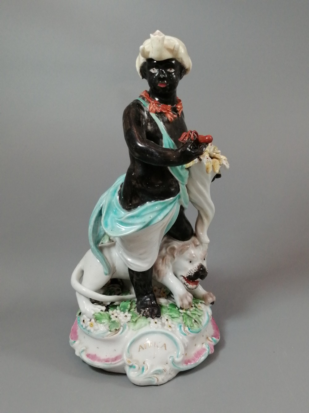 An 18th-century Derby soft-paste porcelain figurine depicting an allegorical figure of Africa wearing an elephant scalp headdress beside a crouching lion on a rococo scroll base. View VIII