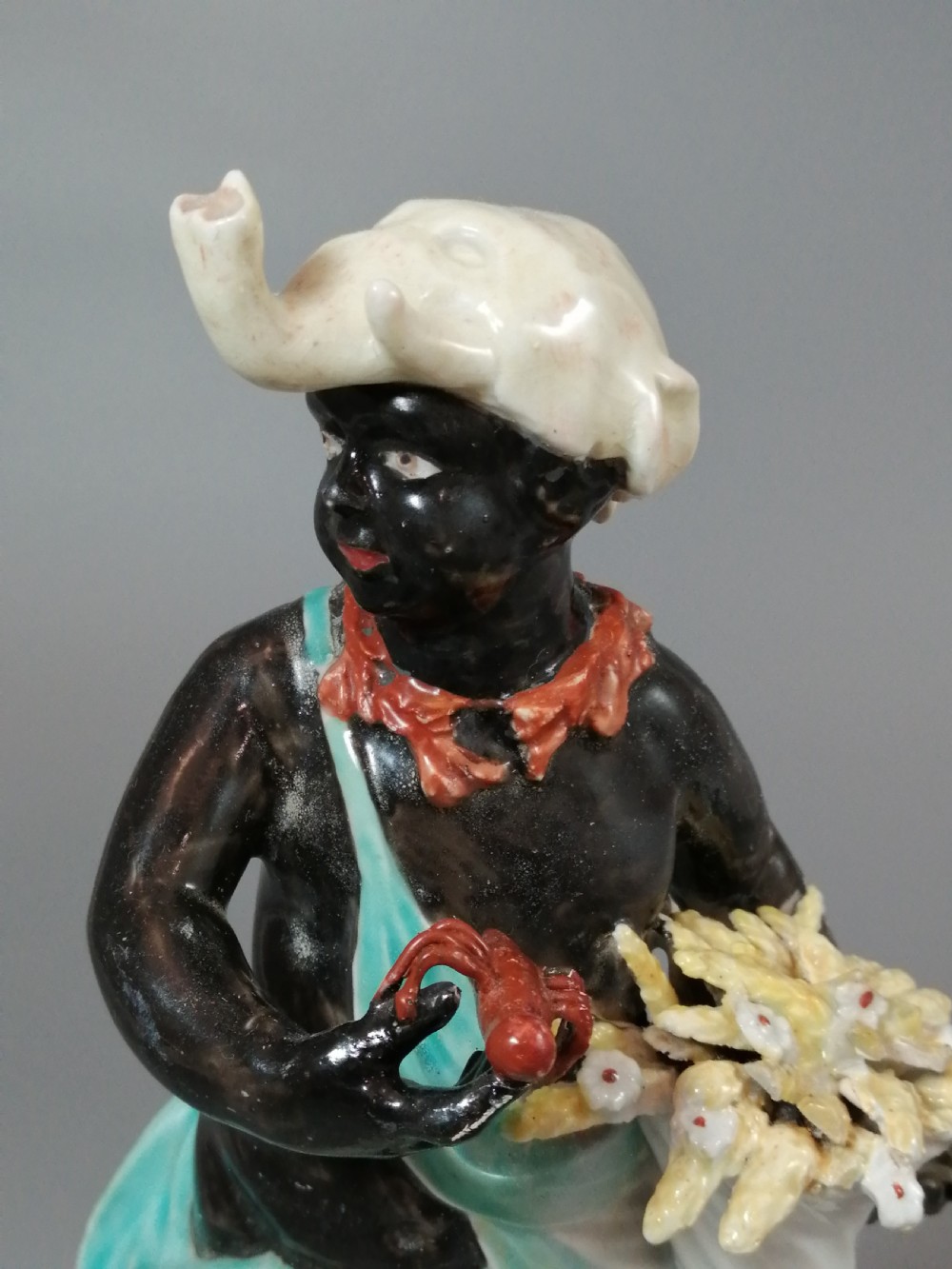 An 18th-century Derby soft-paste porcelain figurine depicting an allegorical figure of Africa wearing an elephant scalp headdress beside a crouching lion on a rococo scroll base. View IX