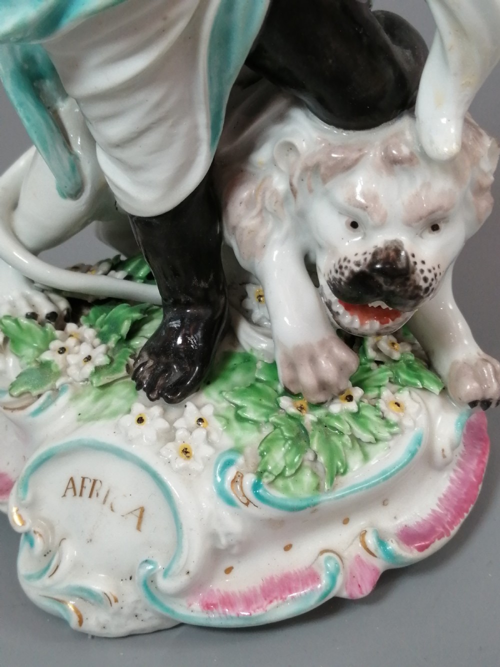 An 18th-century Derby soft-paste porcelain figurine depicting an allegorical figure of Africa wearing an elephant scalp headdress beside a crouching lion on a rococo scroll base. View X
