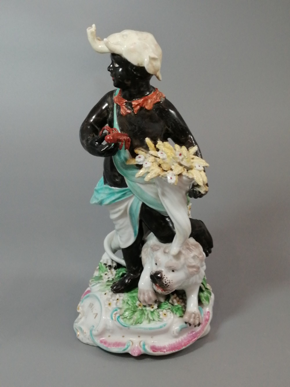 An 18th-century Derby soft-paste porcelain figurine depicting an allegorical figure of Africa wearing an elephant scalp headdress beside a crouching lion on a rococo scroll base. View XI