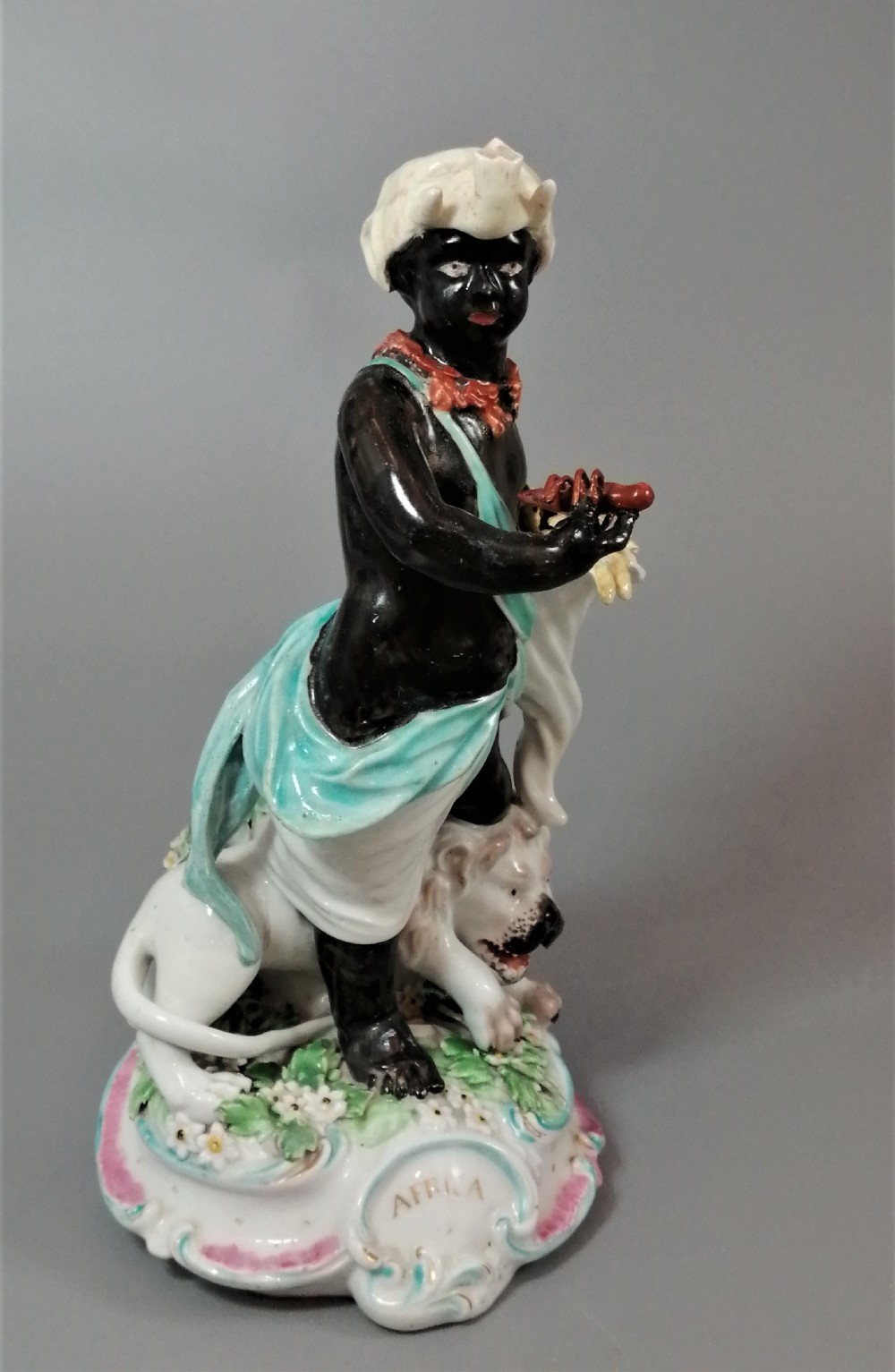 An 18th-century Derby soft-paste porcelain figurine depicting an allegorical figure of Africa wearing an elephant scalp headdress beside a crouching lion on a rococo scroll base. View XII