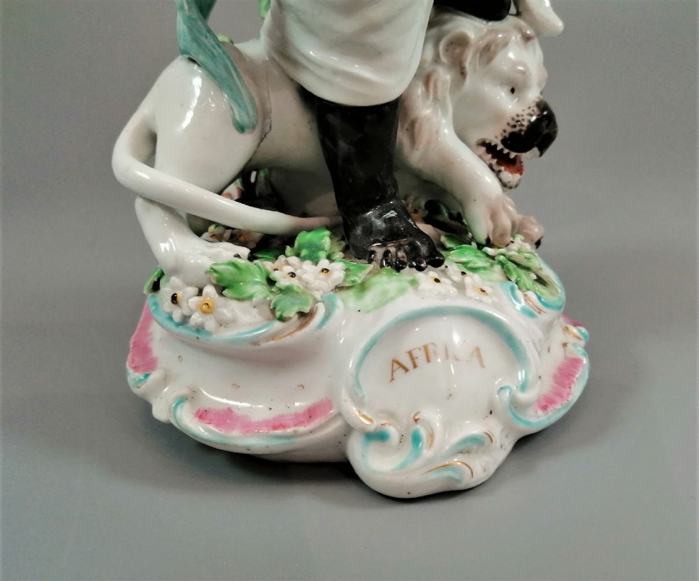 An 18th-century Derby soft-paste porcelain figurine depicting an allegorical figure of Africa wearing an elephant scalp headdress beside a crouching lion on a rococo scroll base. View XIII