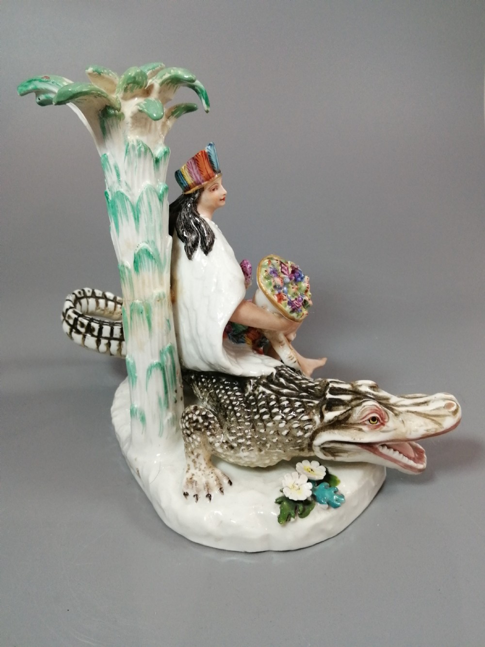 An 18th-century Chelsea polychrome porcelain statuette depicting an allegorical female figure seated on an alligator beside a palm tree. View VI