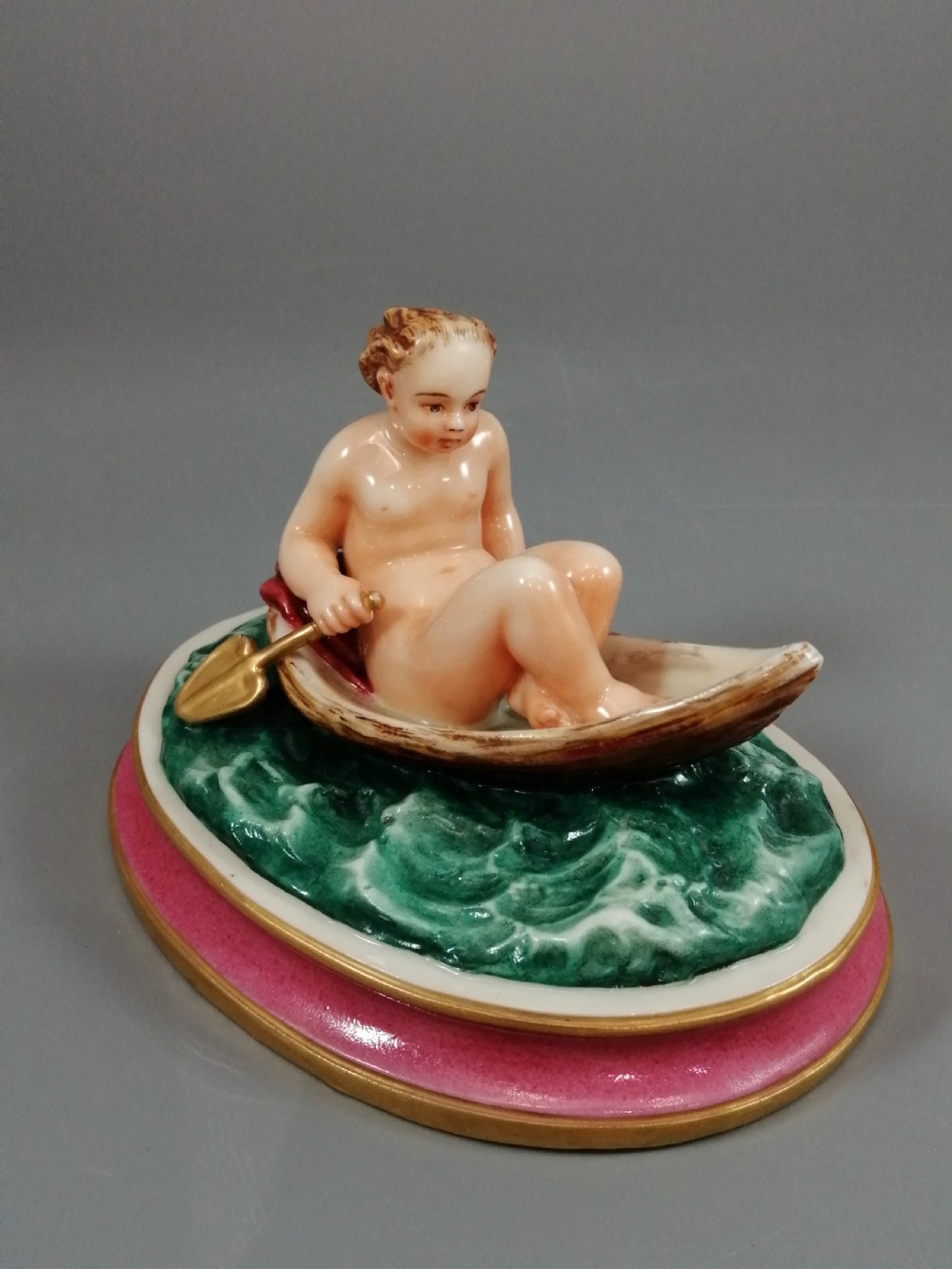 A 19th-century majolica ceramic figurine depicting a figure seated in a small boat over green waves, mounted on a pink glazed oval base with gilded borders.