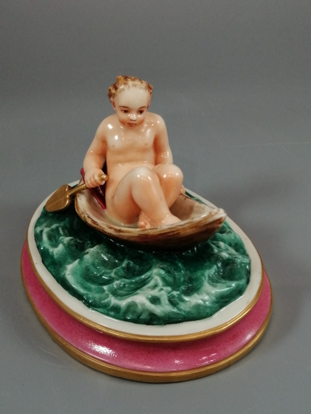 A 19th-century majolica ceramic figurine depicting a figure seated in a small boat over green waves, mounted on a pink glazed oval base with gilded borders. View II