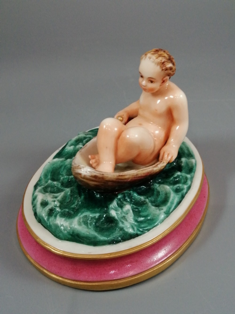 A 19th-century majolica ceramic figurine depicting a figure seated in a small boat over green waves, mounted on a pink glazed oval base with gilded borders. View III