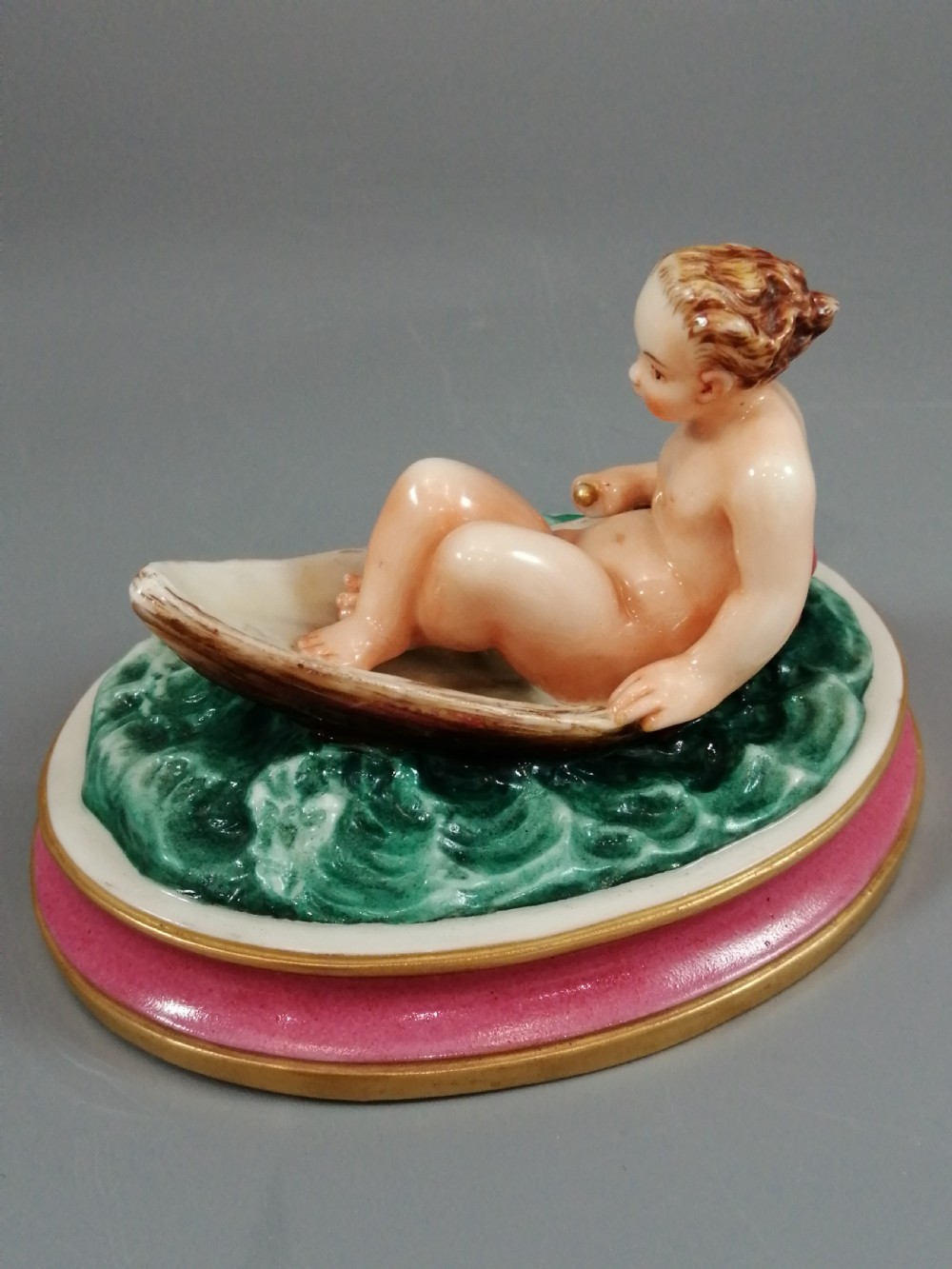 A 19th-century majolica ceramic figurine depicting a figure seated in a small boat over green waves, mounted on a pink glazed oval base with gilded borders. View IV