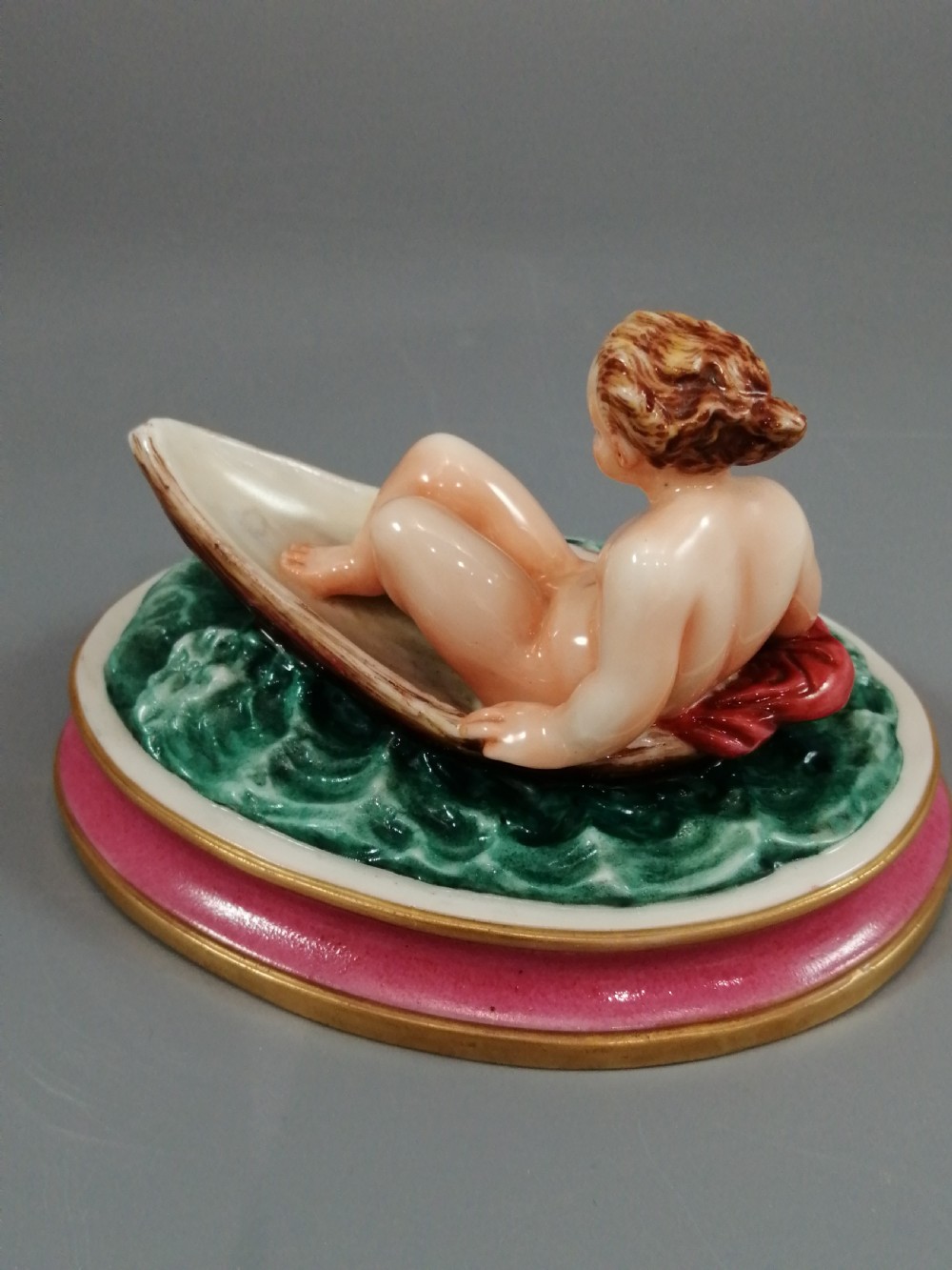 A 19th-century majolica ceramic figurine depicting a figure seated in a small boat over green waves, mounted on a pink glazed oval base with gilded borders. View V