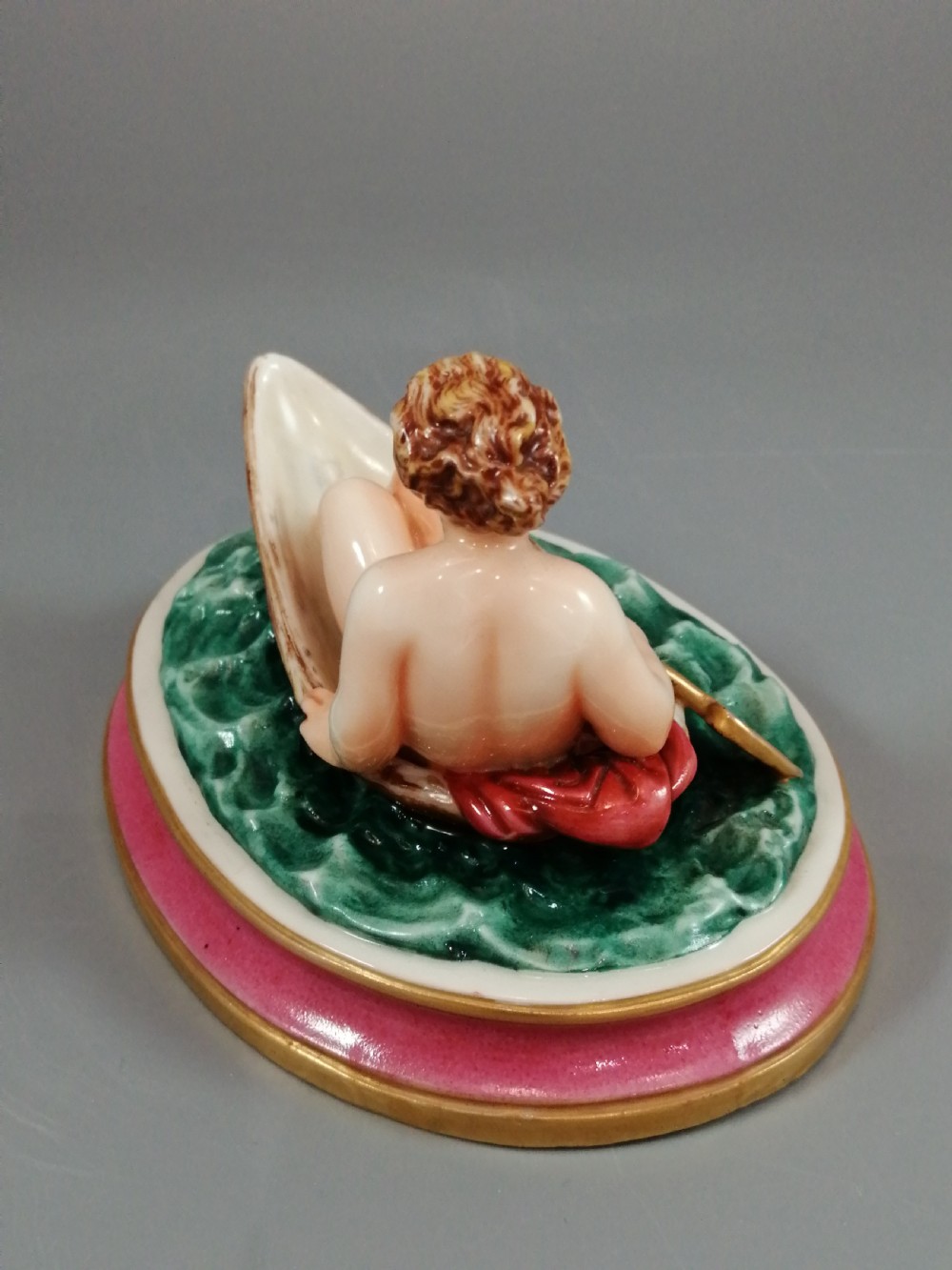 A 19th-century majolica ceramic figurine depicting a figure seated in a small boat over green waves, mounted on a pink glazed oval base with gilded borders. View VI