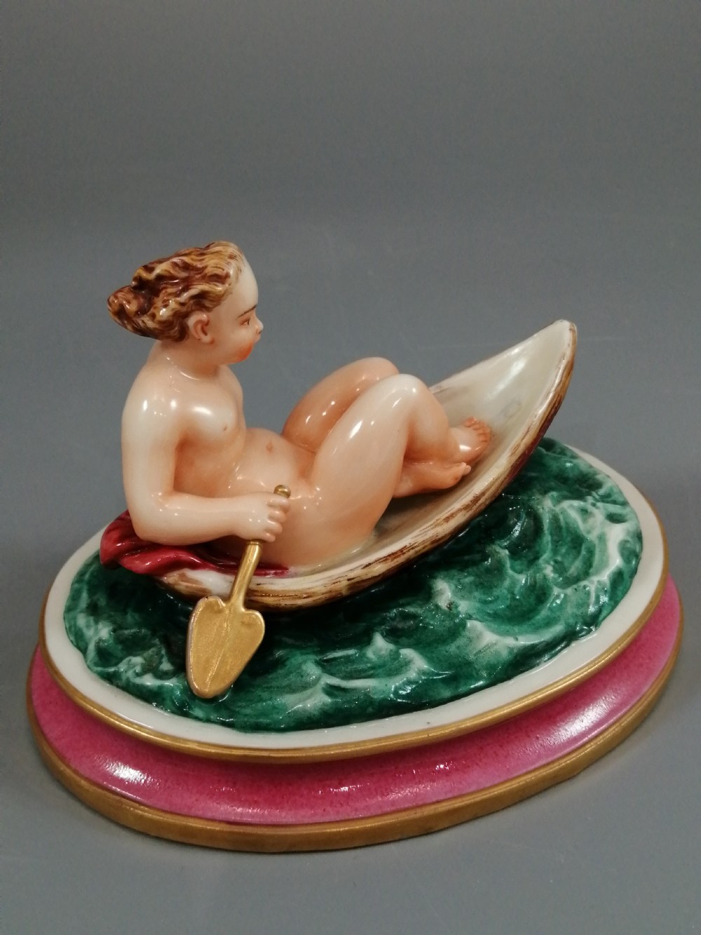 A 19th-century majolica ceramic figurine depicting a figure seated in a small boat over green waves, mounted on a pink glazed oval base with gilded borders. View VIII