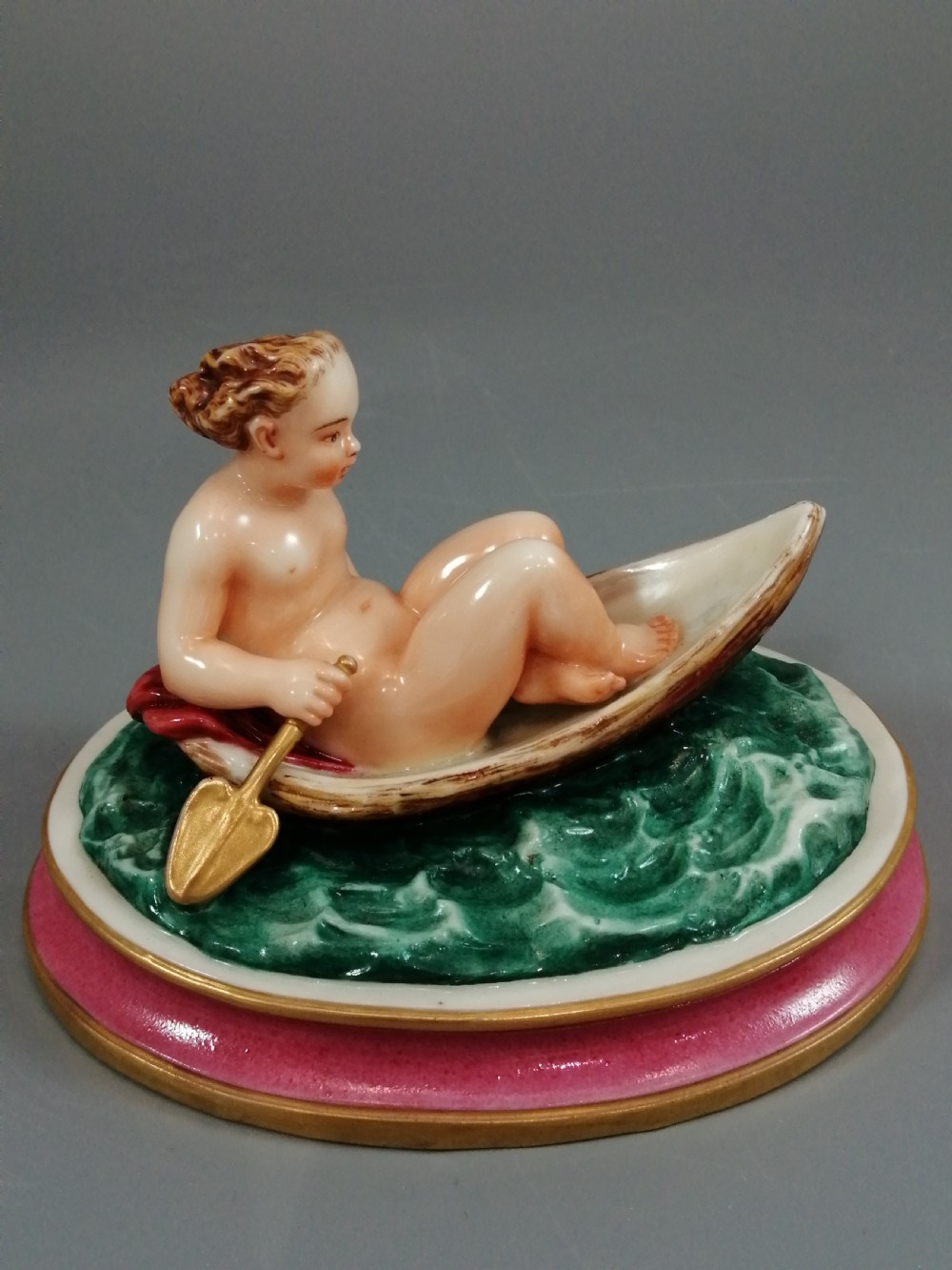 A 19th-century majolica ceramic figurine depicting a figure seated in a small boat over green waves, mounted on a pink glazed oval base with gilded borders. View IX