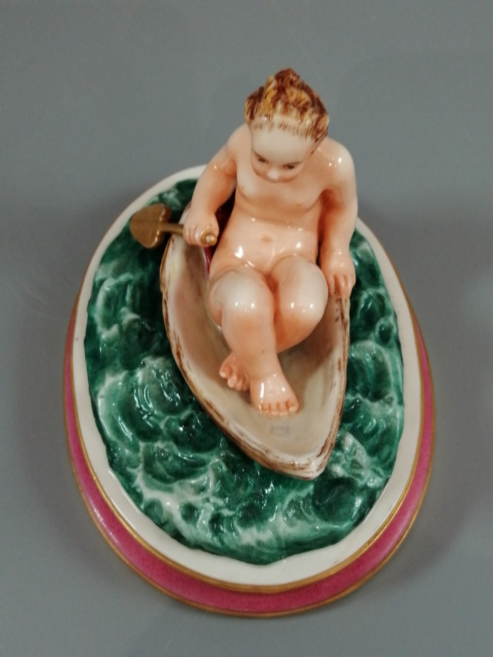 A 19th-century majolica ceramic figurine depicting a figure seated in a small boat over green waves, mounted on a pink glazed oval base with gilded borders. View XI