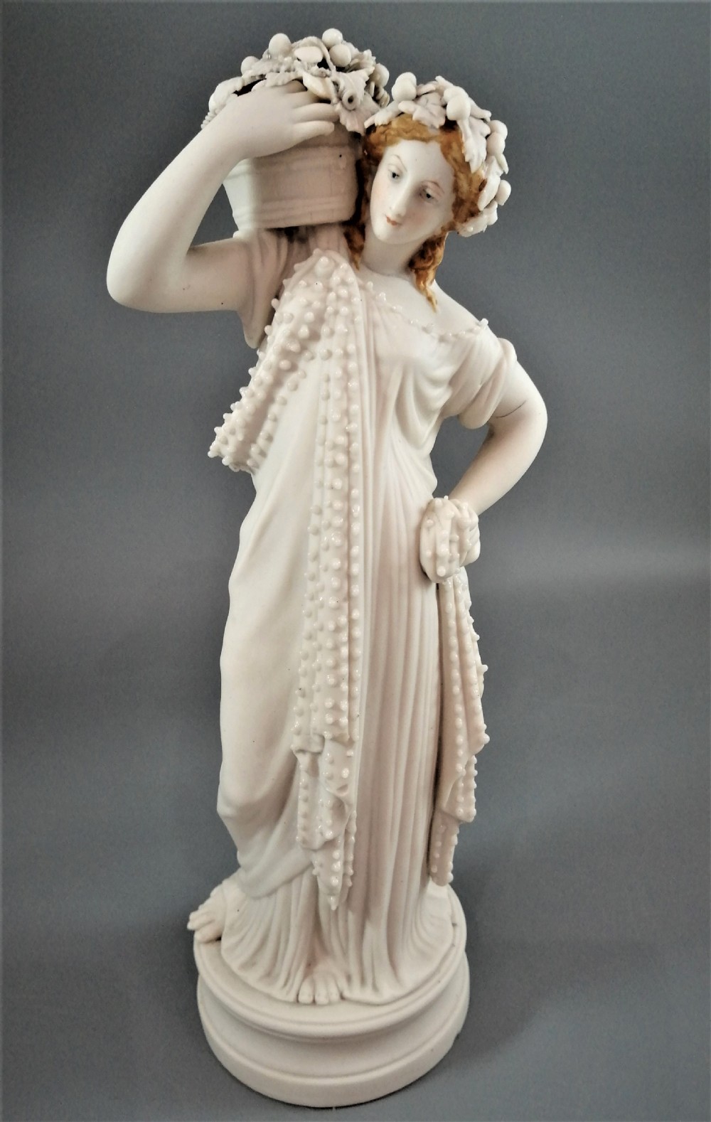 A nineteenth-century Victorian Parian ware figurine depicting a classically draped maiden holding a fruit basket on her shoulder.