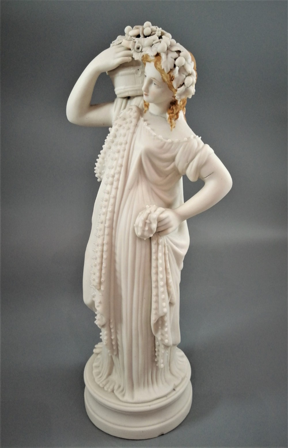 A nineteenth-century Victorian Parian ware figurine depicting a classically draped maiden holding a fruit basket on her shoulder. View II