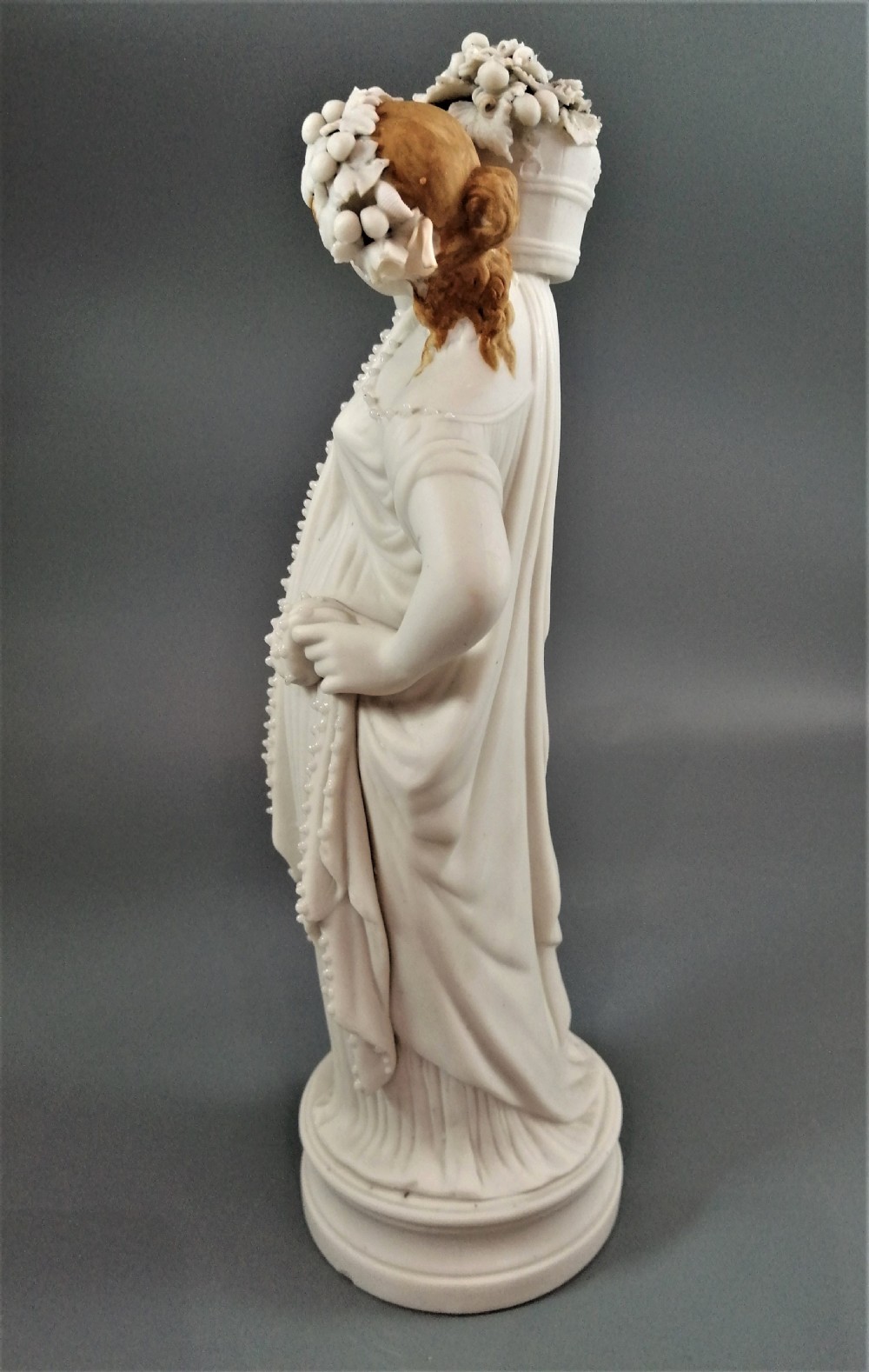 A nineteenth-century Victorian Parian ware figurine depicting a classically draped maiden holding a fruit basket on her shoulder. View III