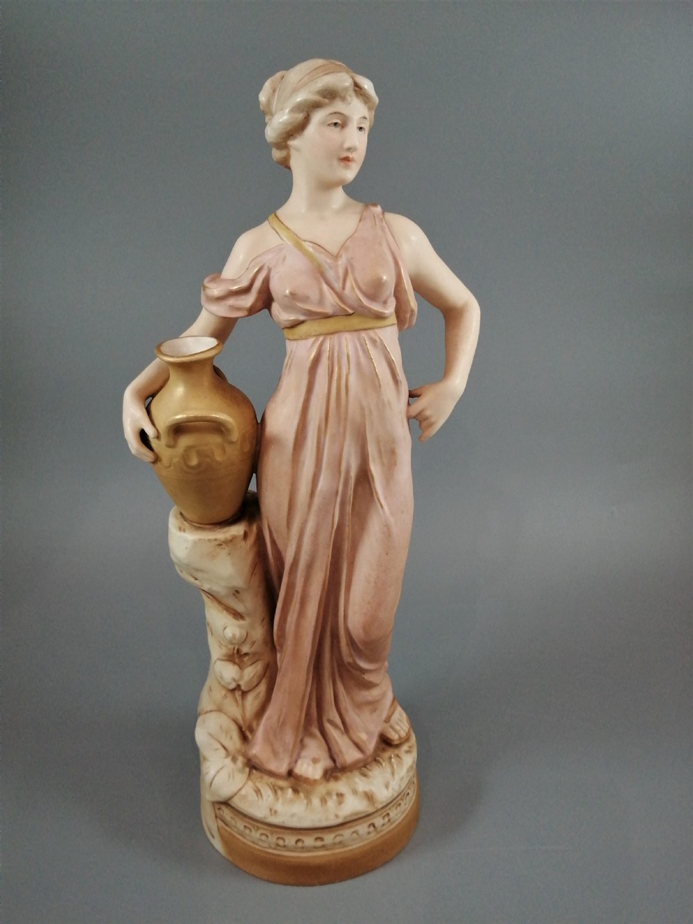 A Royal Dux porcelain statuette of a classical maiden in a draped dusty pink gown holding an earthenware jug.