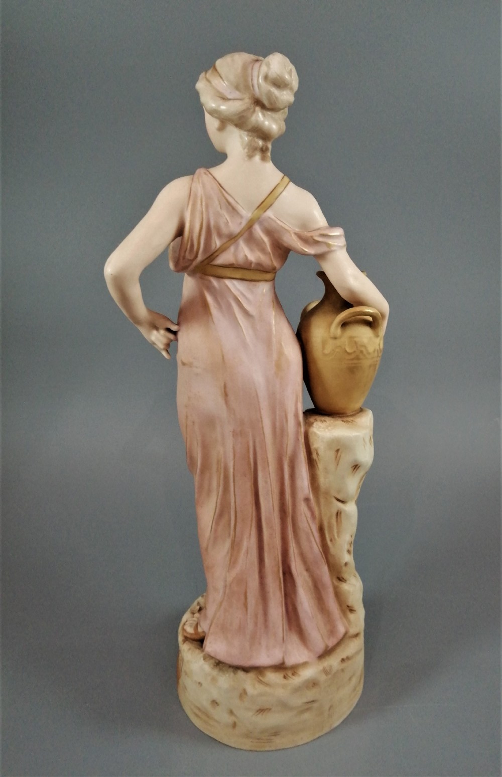 A Royal Dux porcelain statuette of a classical maiden in a draped dusty pink gown holding an earthenware jug. View IV