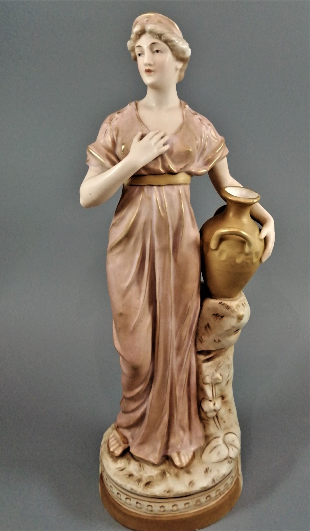 An antique Royal Dux porcelain figurine depicting a woman in a draped pink gown holding a gilded water jug.