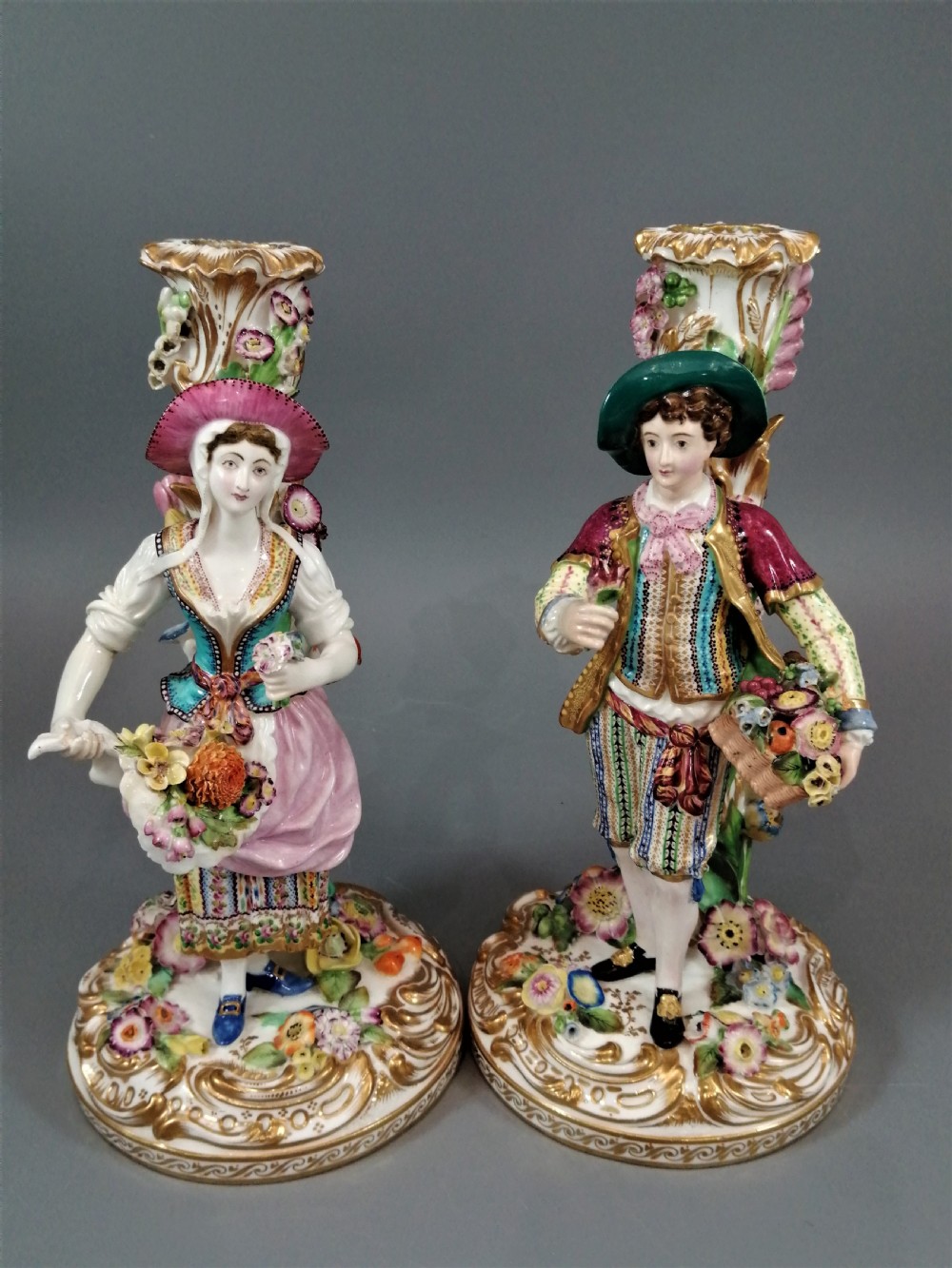 A pair of hand-painted Minton porcelain figural candlesticks featuring a male and female figure in colourful traditional dress with floral motifs and gilded scroll bases.