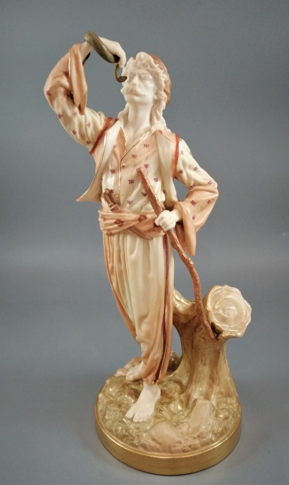 A nineteenth-century Royal Worcester porcelain figurine depicting a standing snake charmer holding a snake aloft beside a tree stump base.