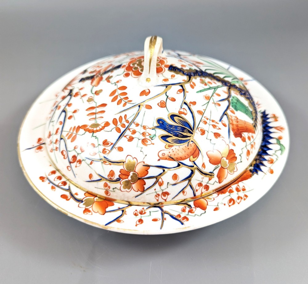 A circular antique porcelain butter dish with stand, decorated with hand-painted Japanese Kakiemon style quail and floral motifs in iron-red, blue, and gold.