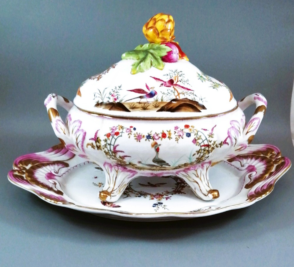An antique continental porcelain soup tureen with a fruit finial lid, dual handles, and matching underplate, decorated with hand-painted birds and gilded scrollwork.