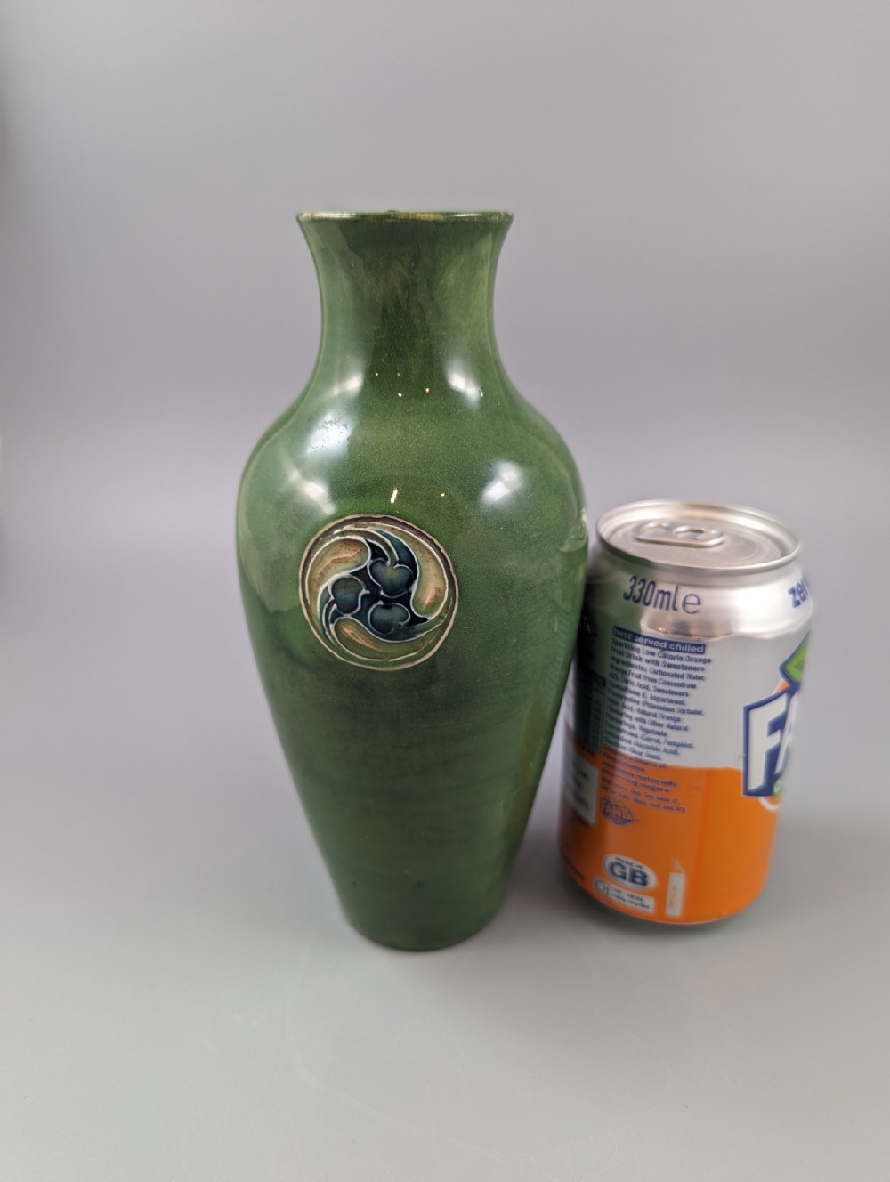 A green glazed ceramic vase with a flared neck and a central circular slip-trailed medallion featuring a stylized motif in blue, green, and cream. View X