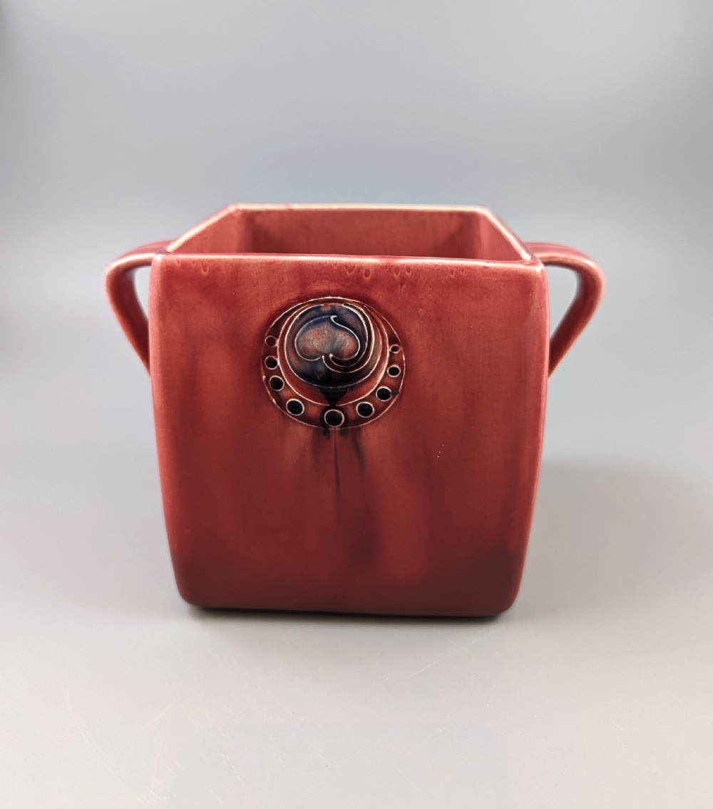 A square red glazed ceramic vessel with twin loop handles and a central circular motif on the front surface.