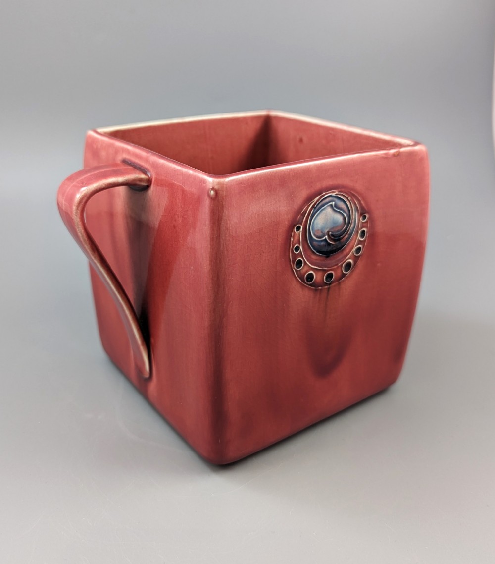 A square red glazed ceramic vessel with twin loop handles and a central circular motif on the front surface. View III