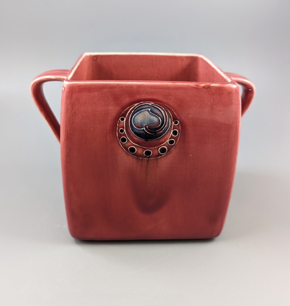 A square red glazed ceramic vessel with twin loop handles and a central circular motif on the front surface. View IV