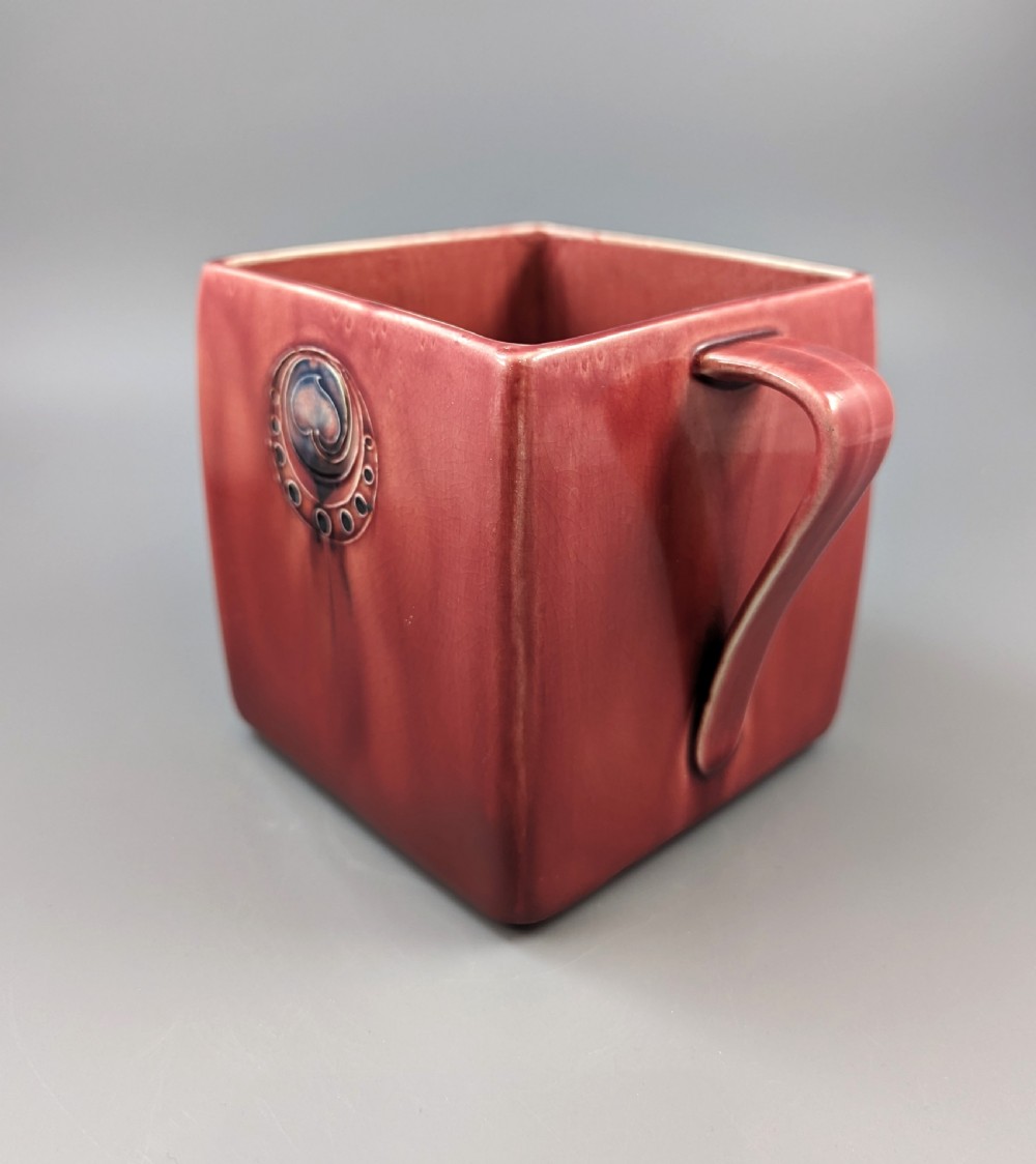 A square red glazed ceramic vessel with twin loop handles and a central circular motif on the front surface. View V