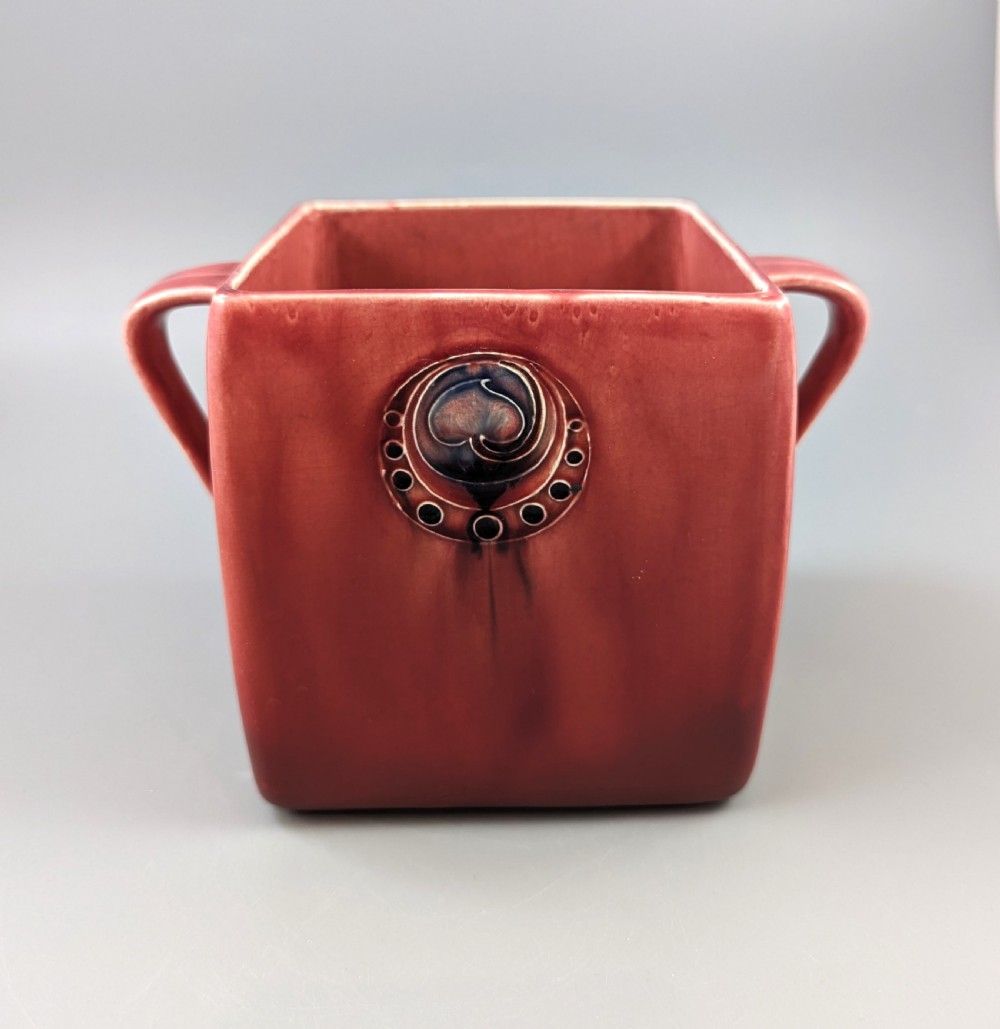 A square red glazed ceramic vessel with twin loop handles and a central circular motif on the front surface. View VI